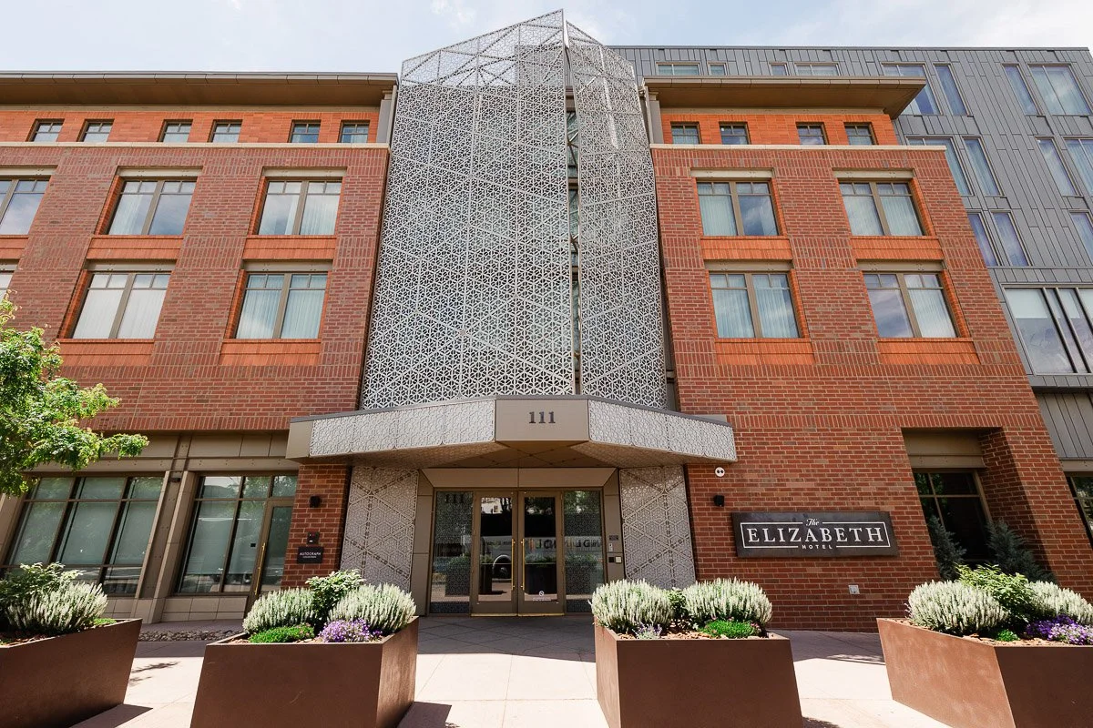 Facade of a modern brick building with geometric metal design over entrance, labeled "Elizabeth." Potted plants line the entrance, giving a welcoming feel.