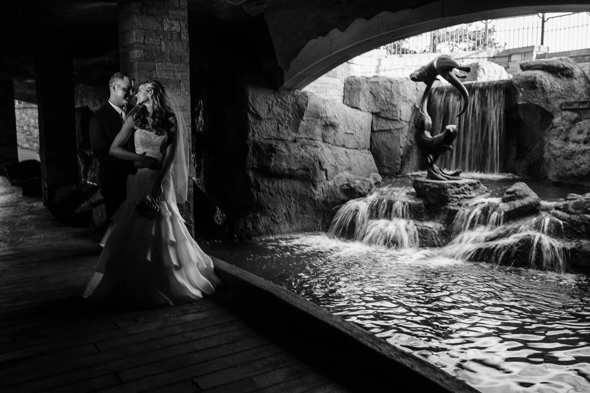 A bride and groom embrace under a stone archway beside a waterfall featuring a dolphin statue. The black and white image conveys a romantic, serene mood during a Sanctuary Golf Course wedding in Sedalia, Colorado.