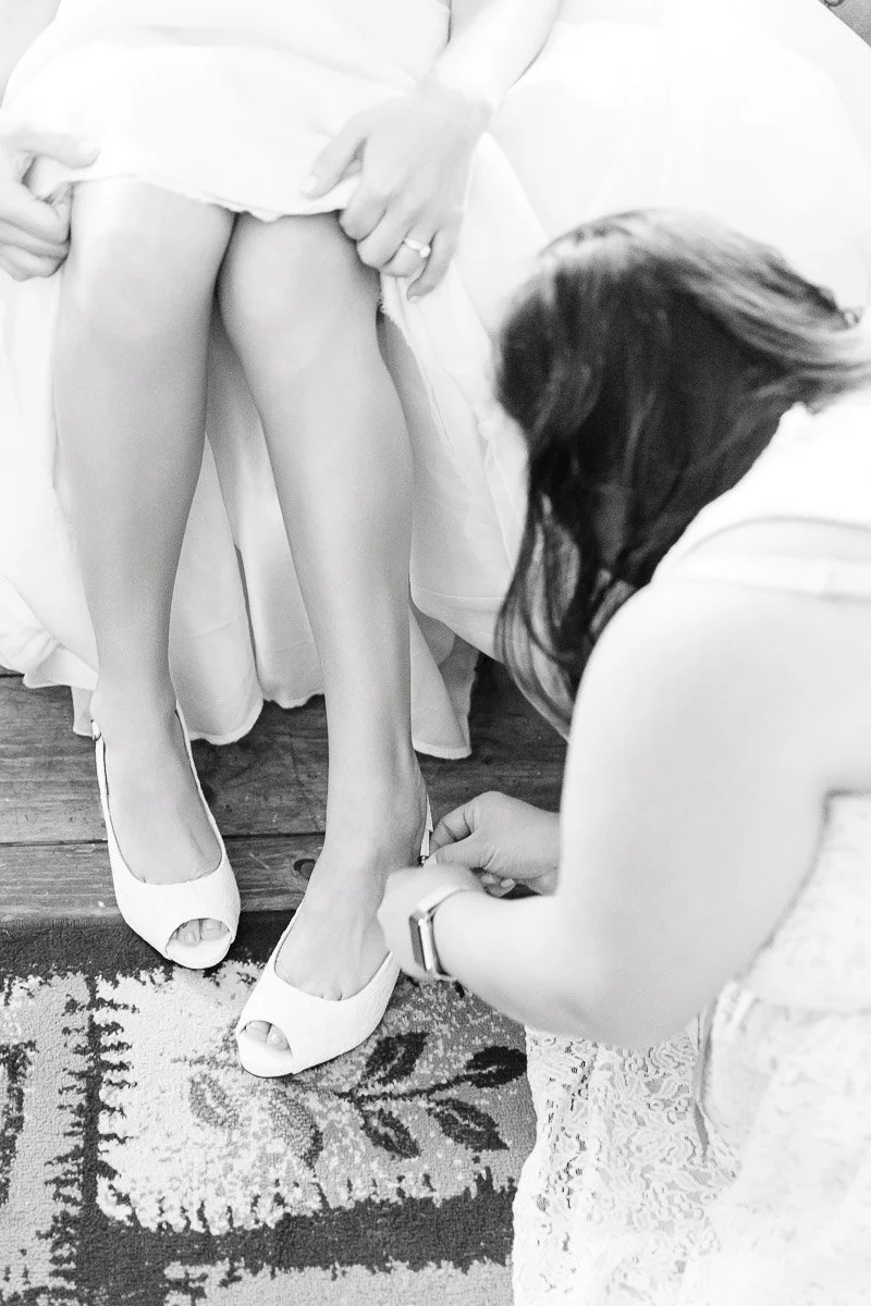 Black and white image of a woman helping another into open-toed heels, emphasizing a tender wedding preparation moment. Elegant, intimate setting.