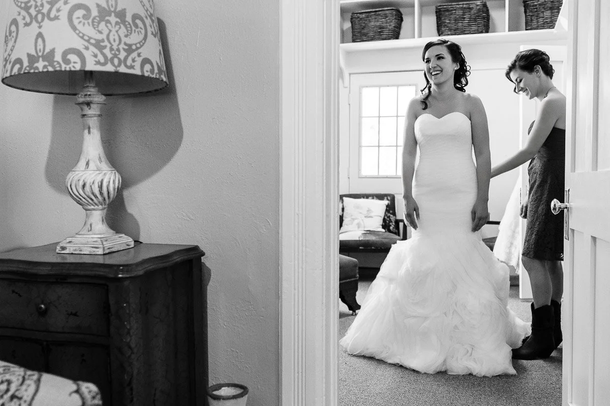 Bride in a white gown smiles as an attendant adjusts her dress in a cozy room. Warm lighting and elegant decor create a joyful, intimate atmosphere.