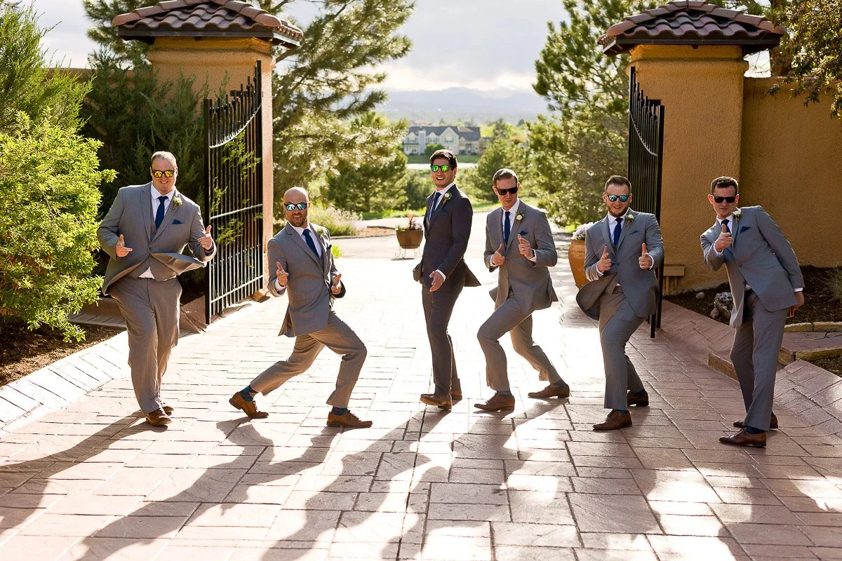Groomsmen in gray suits and blue ties pose playfully with thumbs up under sunny skies, standing on a brick path with greenery and gates behind.