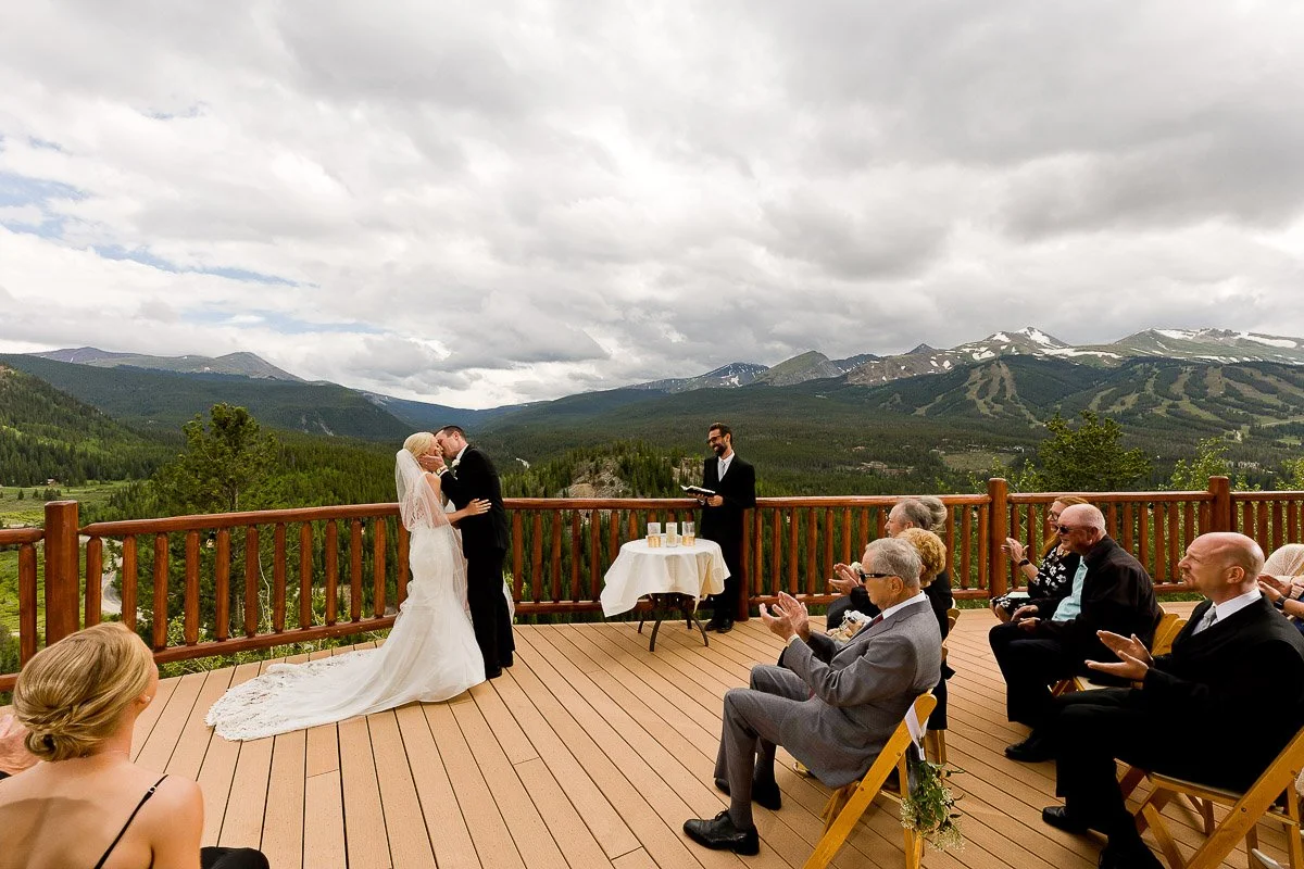 A bride and groom kiss at an outdoor wedding on a wooden deck with mountains in the background. Guests, seated and clapping, witness the ceremony.