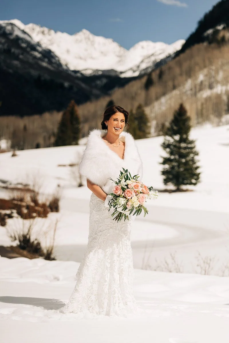 Bride in a white gown and fur stole smiles, holding a pink and white bouquet, standing in a snowy mountain landscape with a bright sky.