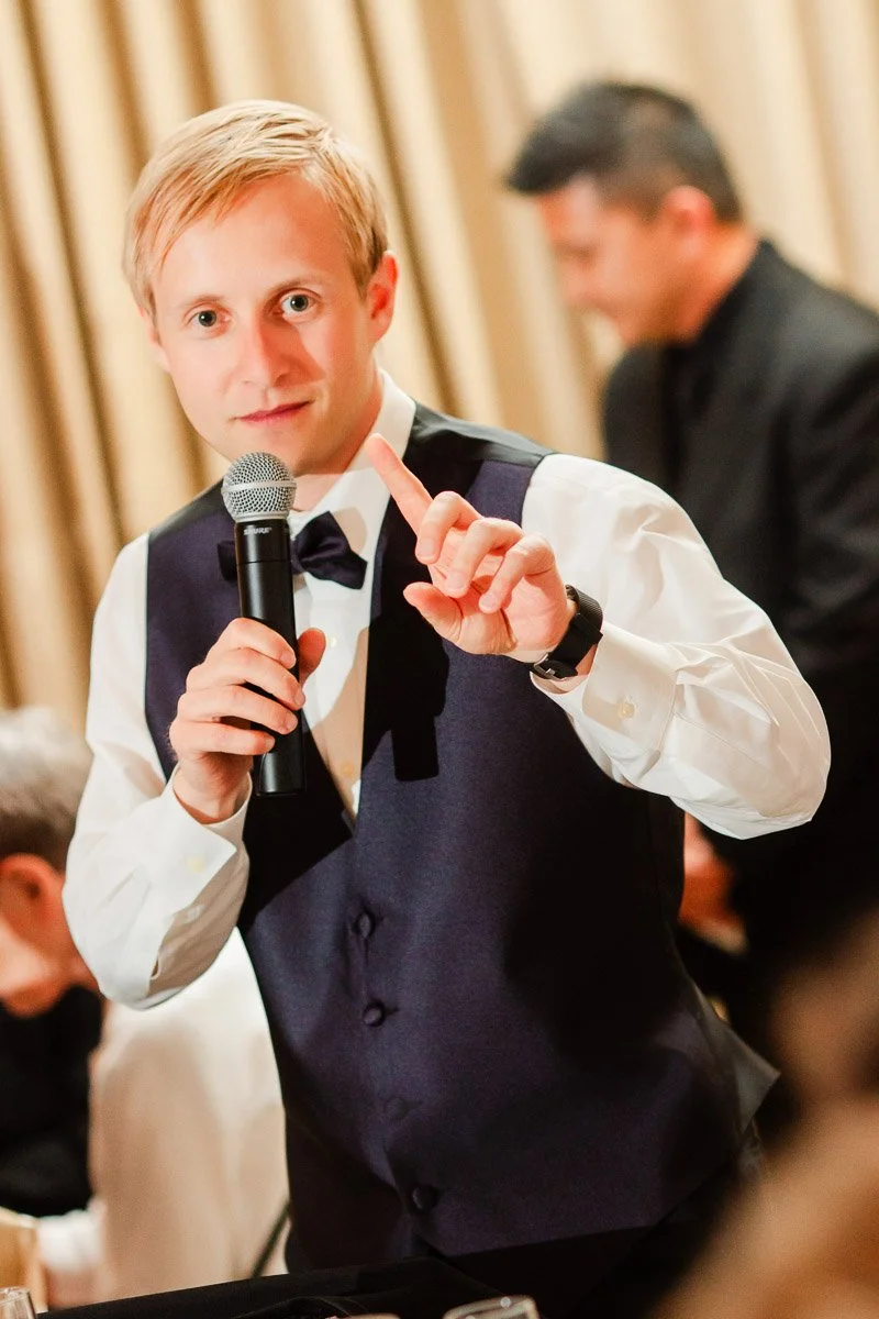 A man in a formal vest and bow tie holds a microphone, gesturing with one finger raised. The setting appears lively, with blurred guests in the background.