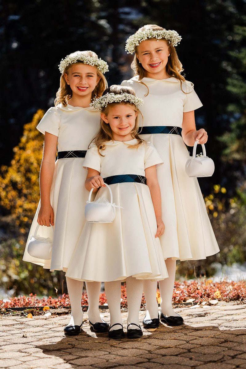Three young girls in matching pale cream dresses and floral crowns stand outdoors on a sunny day. Each holds a small white purse, smiling warmly.