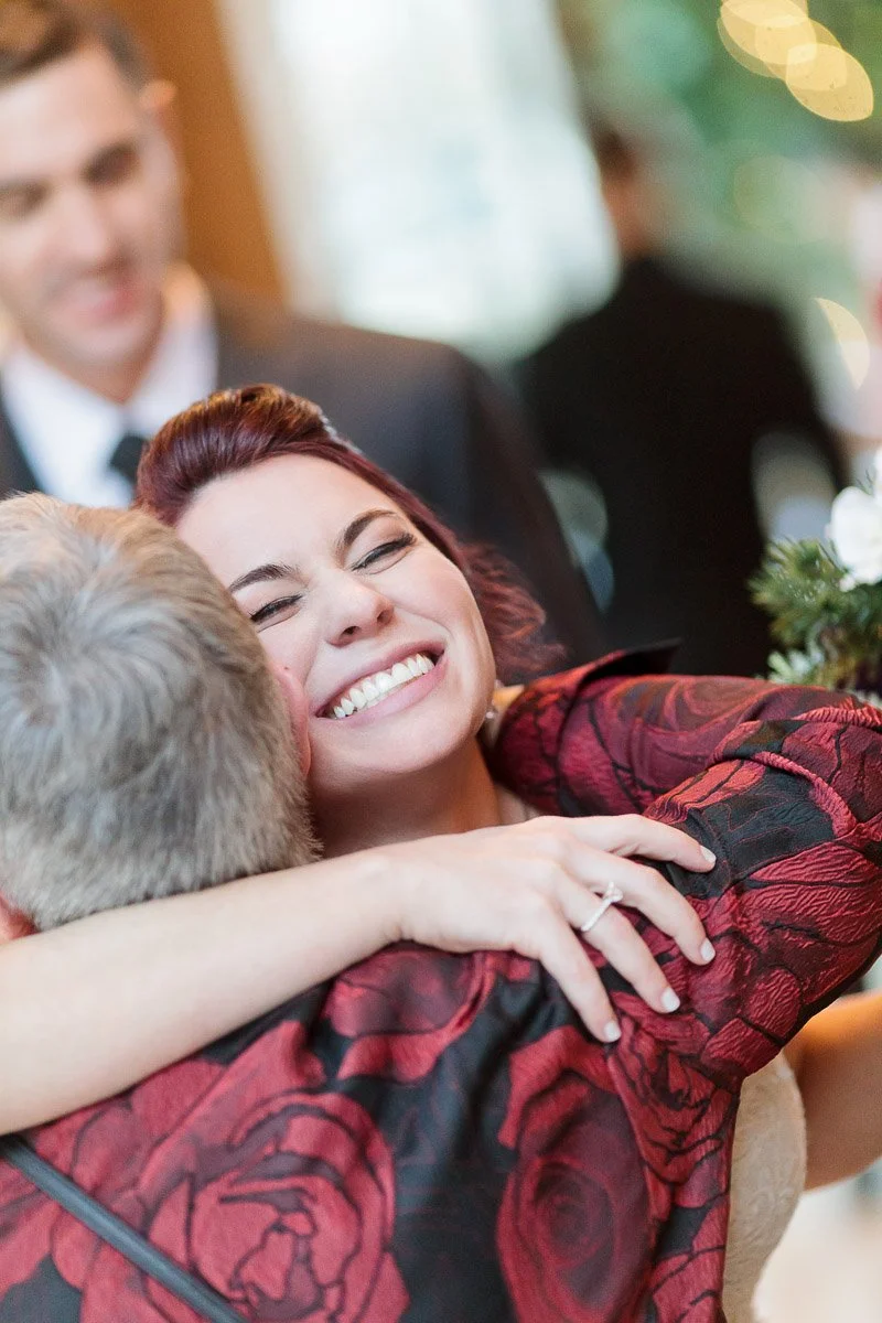 A woman with closed eyes and a wide smile hugs someone joyfully, wearing a red floral-patterned outfit. A man in a suit is blurred in the background.