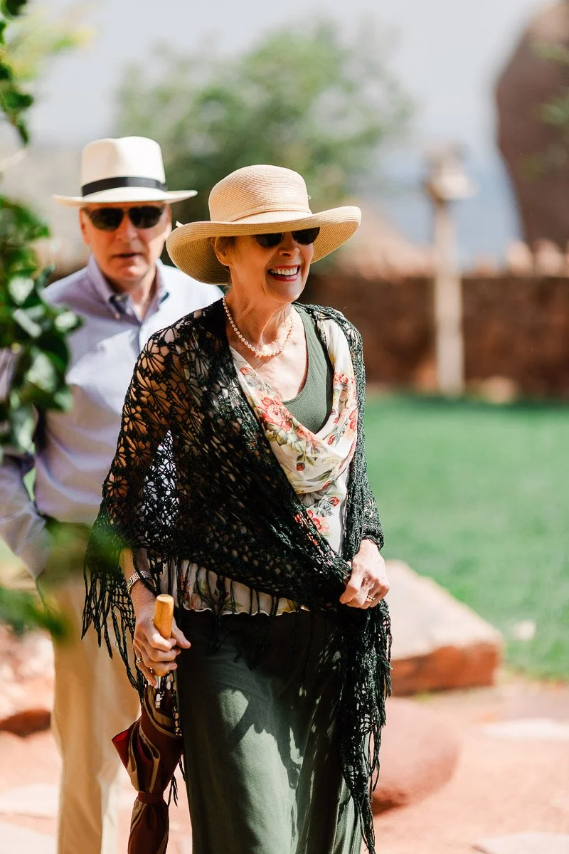 An elderly woman in a sun hat and shawl smiles, holding an umbrella. A man in a white hat follows her. They appear relaxed in a sunny garden.