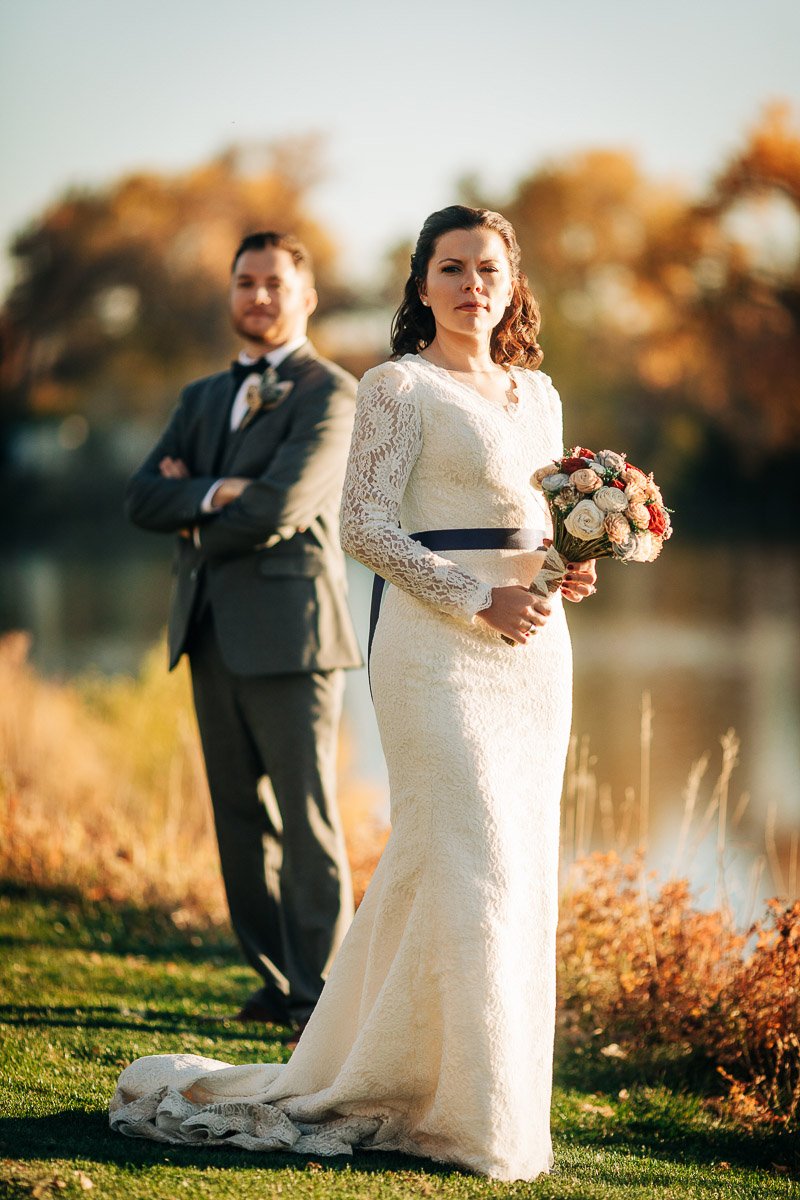 Bride in a lace gown holds a bouquet, standing on grass by a lake during a Wellshire Event Center wedding. Groom in a suit stands behind with arms crossed. Warm autumn tones.