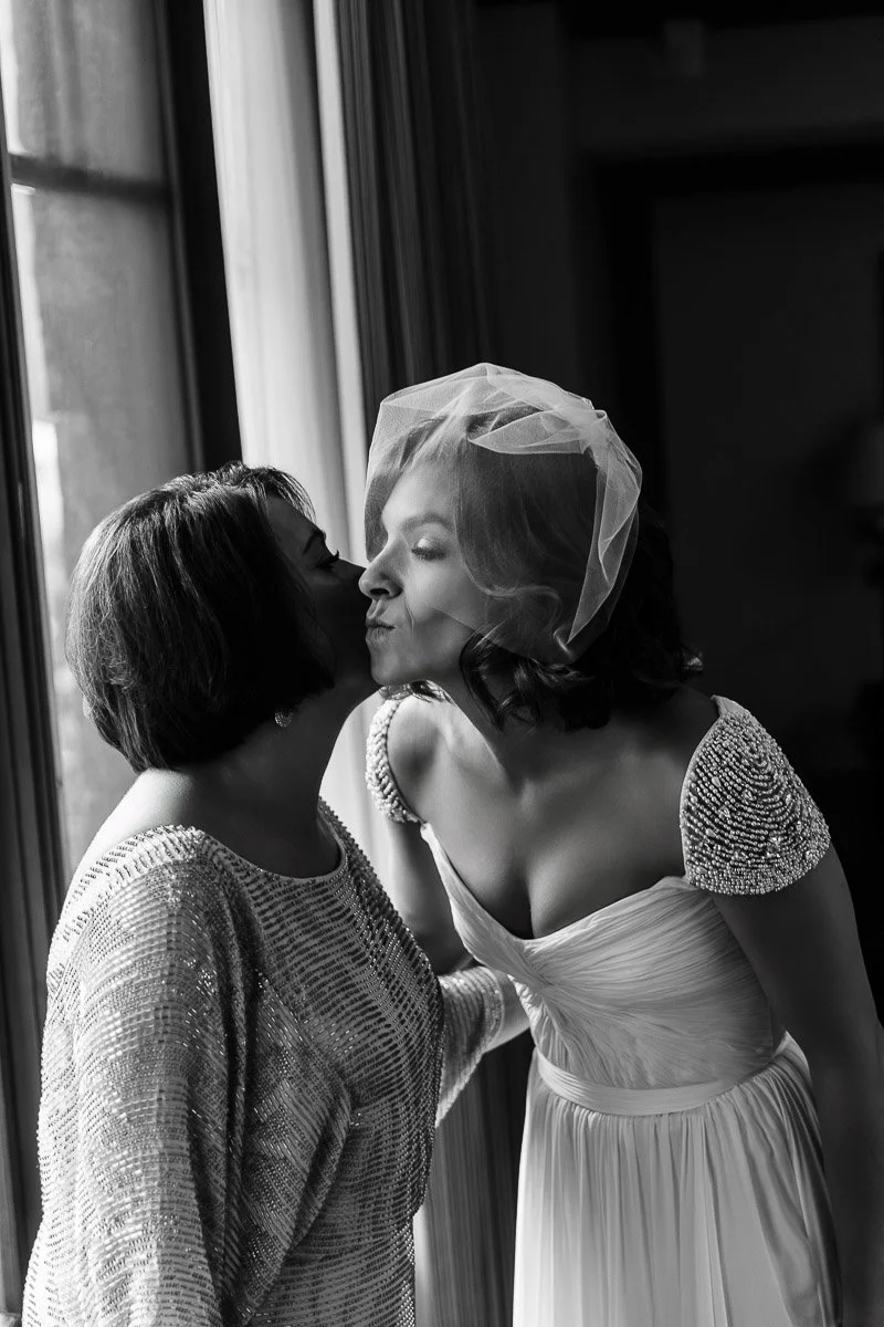 A bride in a wedding dress and veil shares a joyful kiss with a woman by a window. The scene is warm, intimate, and filled with happiness.