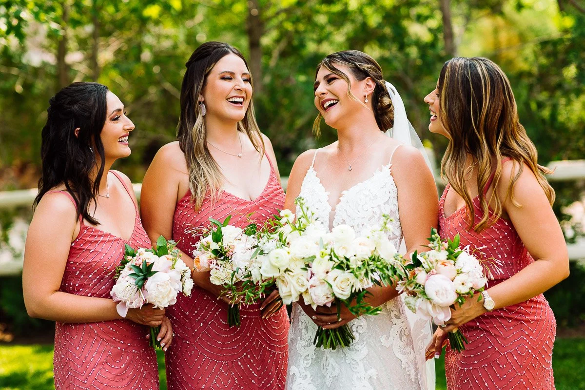 A bride in a white lace gown stands with three bridesmaids in coral dresses, all holding pastel bouquets. They laugh joyfully in a lush outdoor setting.
