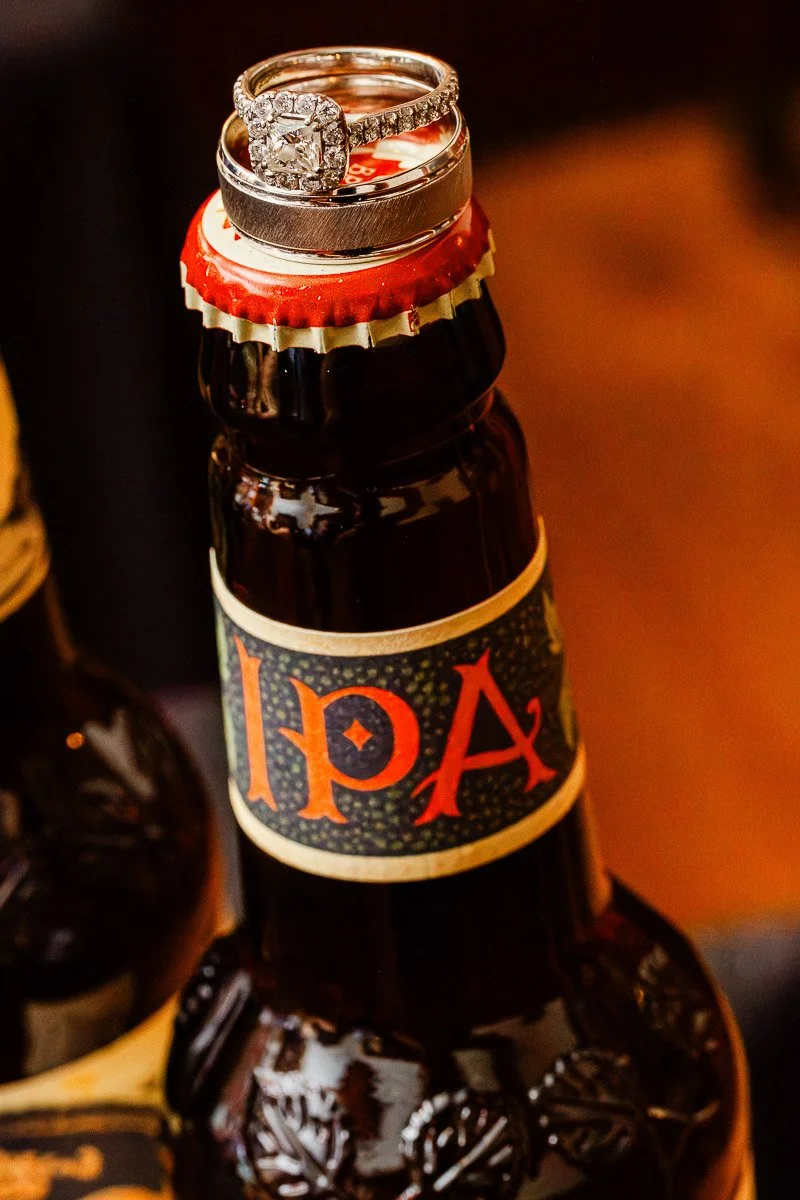 Wedding and engagement rings rest atop an IPA beer bottle with a red cap, highlighting a blend of celebration and casual elegance.