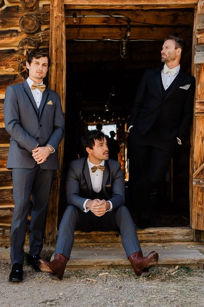 Three men in formal suits with bow ties stand and sit in a rustic wooden doorway. The mood is relaxed, with a mix of casual and formal attire.