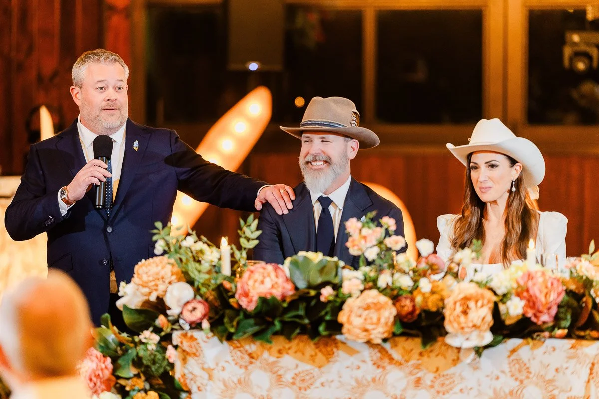 A man in a suit speaks with a microphone, standing beside a smiling bearded man in a hat. A woman in a white hat sits beside them, with a floral table arrangement in front. The setting has a warm, festive atmosphere.