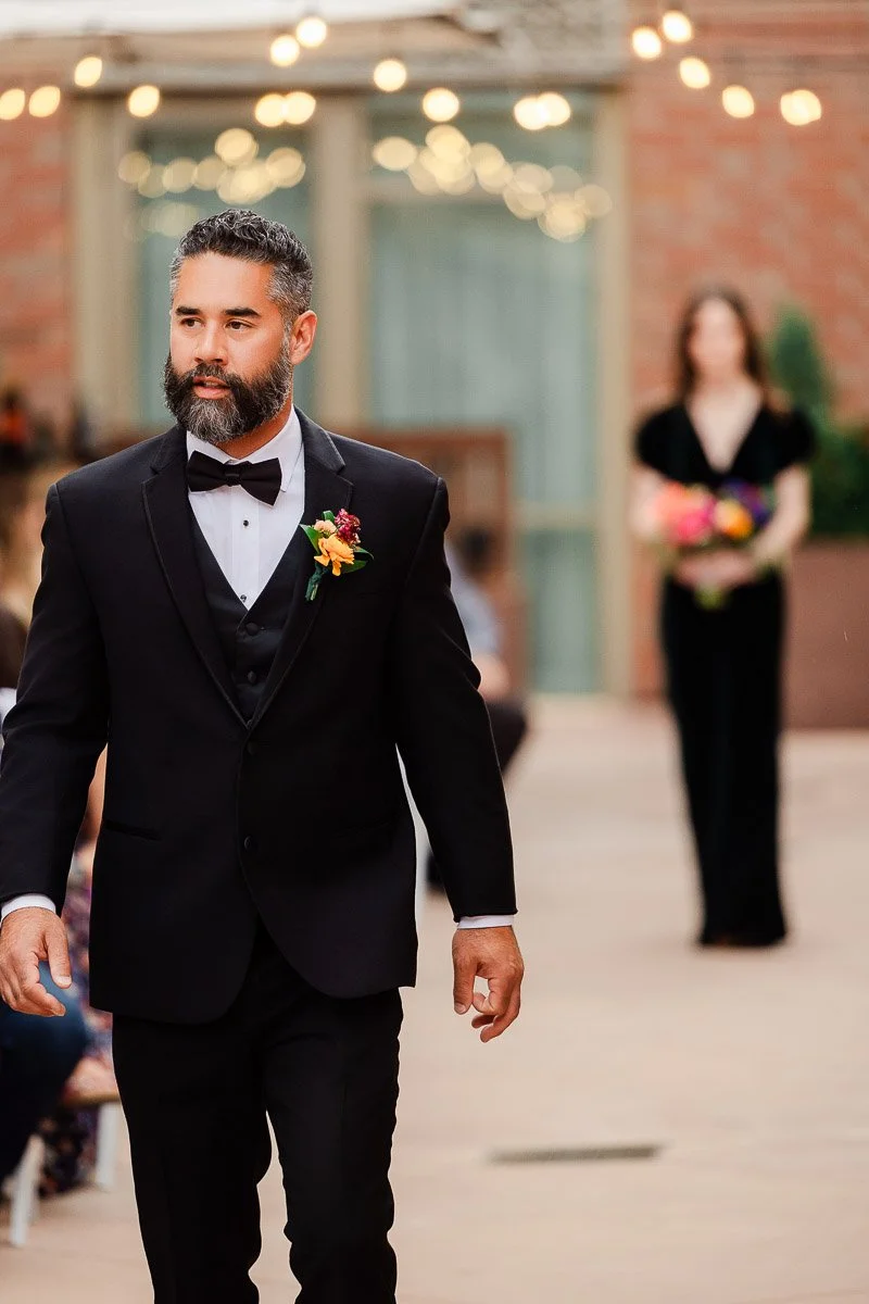 A man in a formal black suit with a bow tie and boutonniere walks confidently outdoors. A woman holding a bouquet stands blurred in the background.