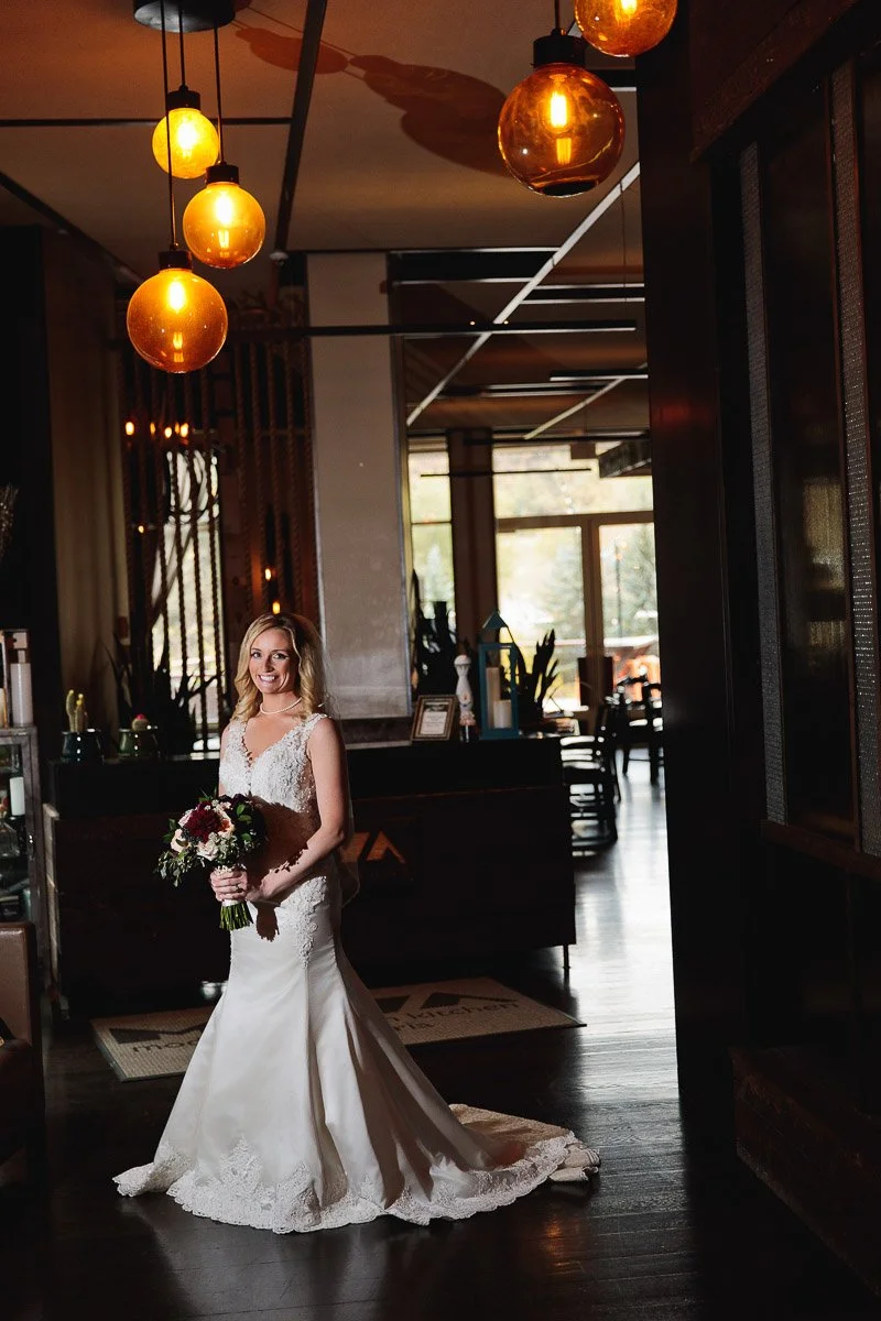 A bride in a lacy white gown stands in an elegant, warmly-lit room with vintage hanging lights. She holds a bouquet, smiling softly during a Westin Riverfront Resort wedding.