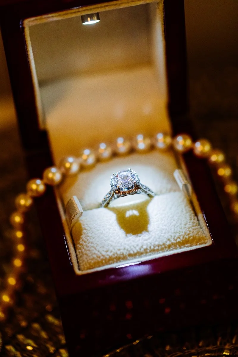 A sparkling diamond ring sits elegantly in a lit, open jewelry box, surrounded by a string of pearls, conveying luxury and sophistication.
