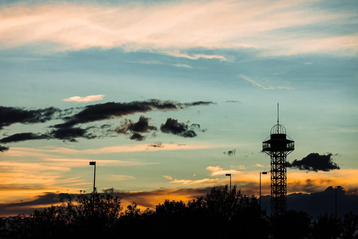 A tall, silhouetted tower stands against a vibrant Denver sunset sky with orange, blue, and gold hues. Wispy clouds add depth above distant trees.