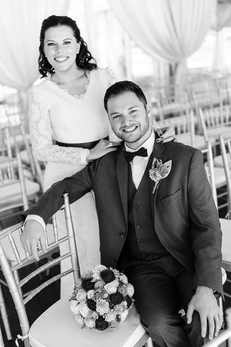 Bride in a lace dress and groom in a suit with a bow tie smile in a bright venue. He holds a bouquet, seated on a chair; joyful and elegant atmosphere.
