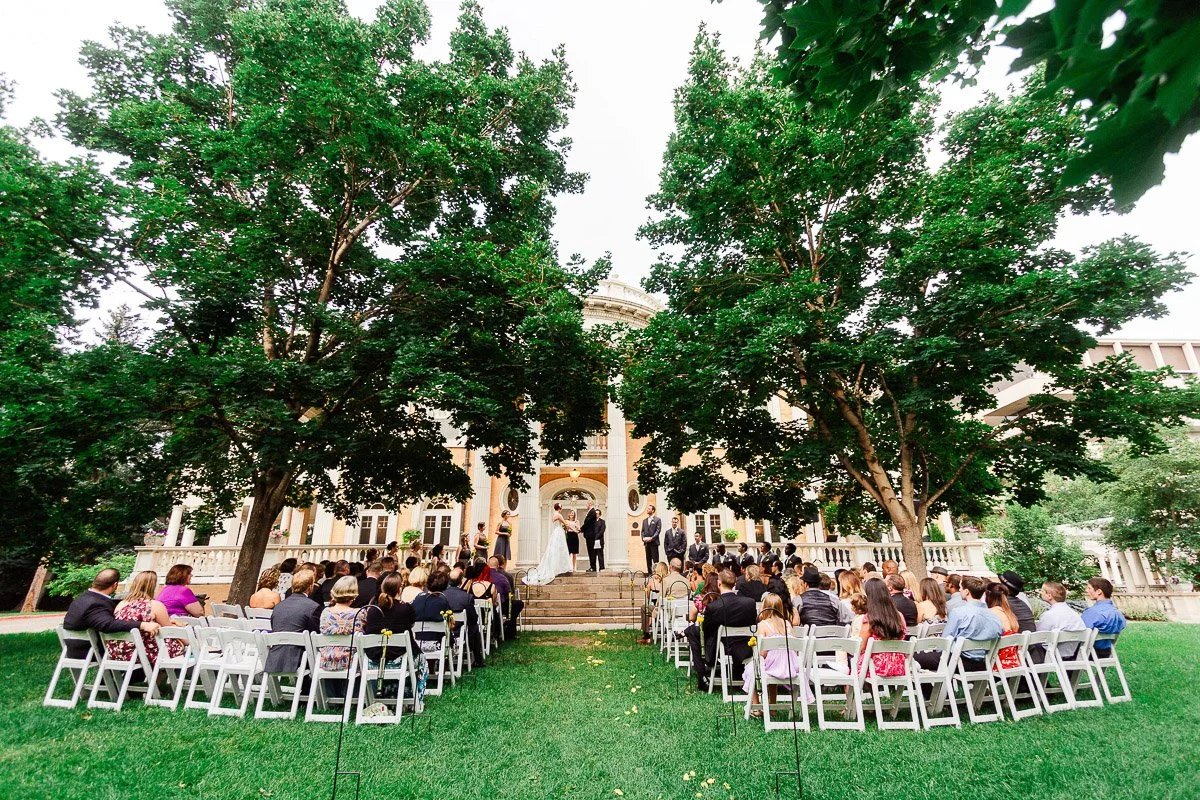 Outdoor GRANT HUMPHREYS MANSION wedding ceremony under two large trees, with guests seated on white chairs facing a couple at an altar in front of a grand building.