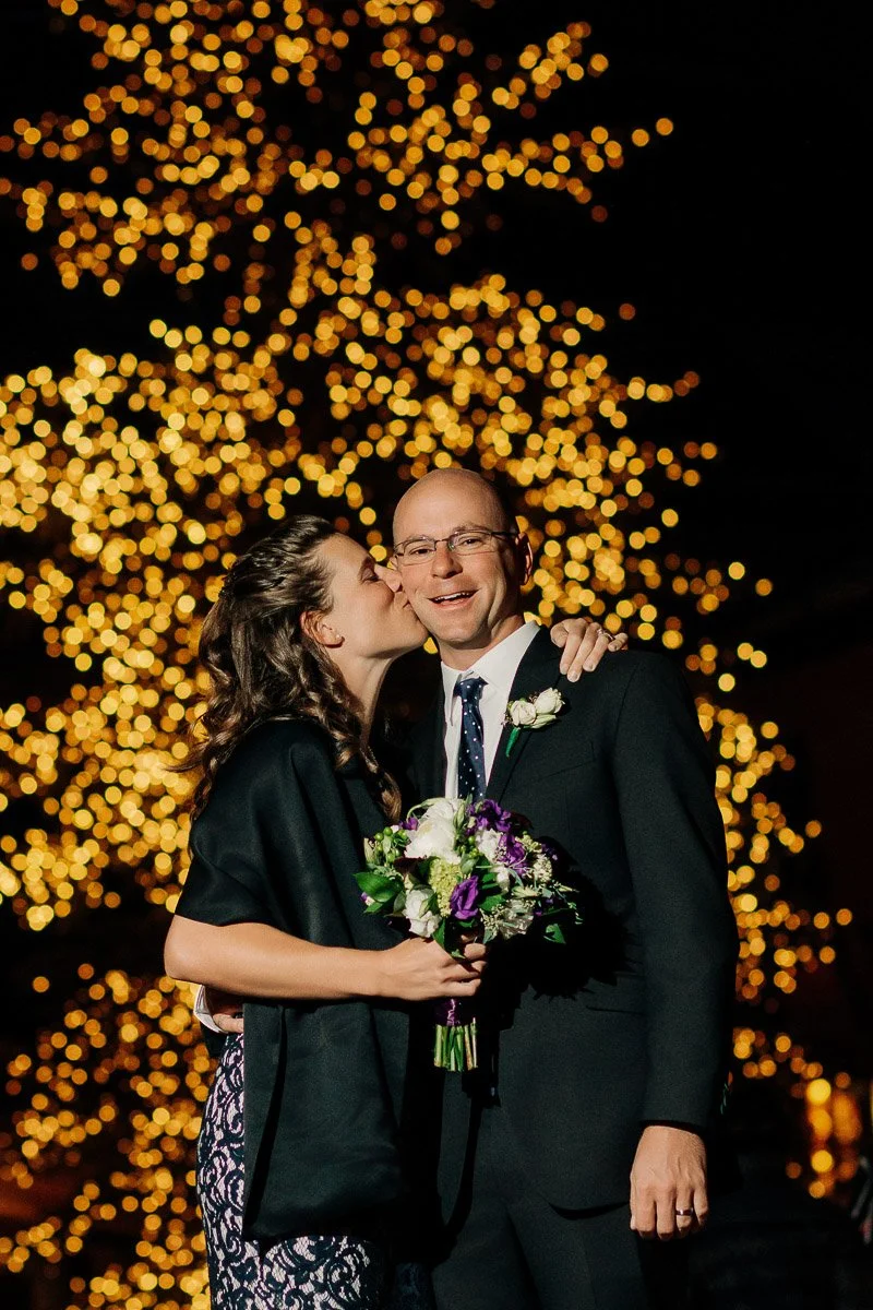 A woman kisses a smiling man on the cheek in front of a glowing backdrop of warm, golden fairy lights. She holds a bouquet, conveying a joyful, celebratory mood.