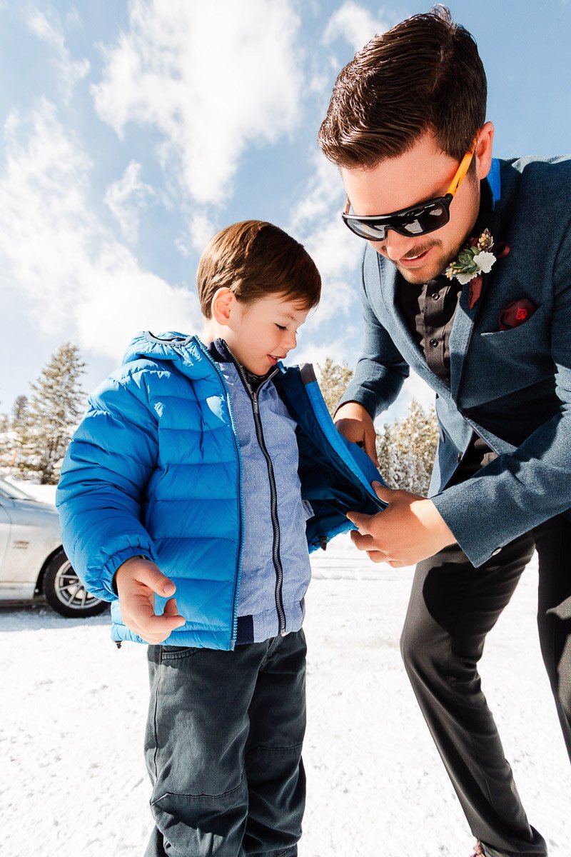 A man in sunglasses helps a child zip up a bright blue jacket in a snowy setting. The scene conveys care and warmth under a sunny sky.