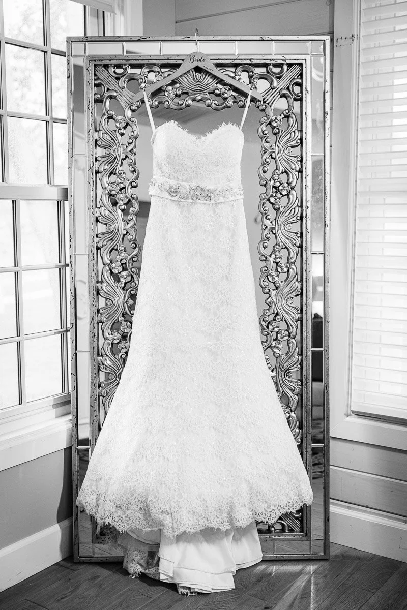 Elegant white lace wedding dress hanging on an ornate mirror frame in a softly lit room. The dress exudes elegance and anticipation.