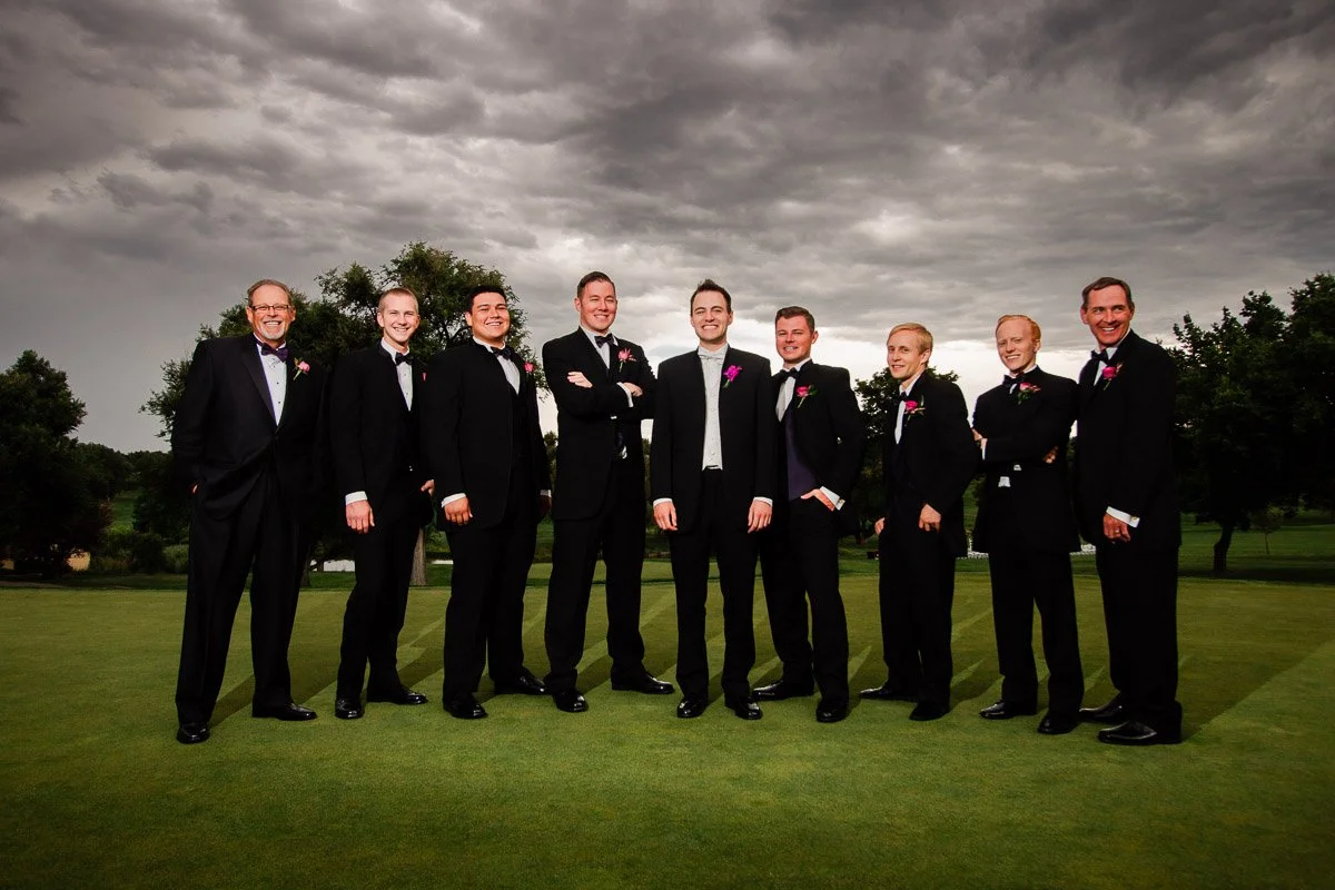 A group of nine men in tuxedos with pink boutonnieres stand on a green field under a cloudy sky, exuding a celebratory and formal atmosphere during a Lakewood Country Club wedding.