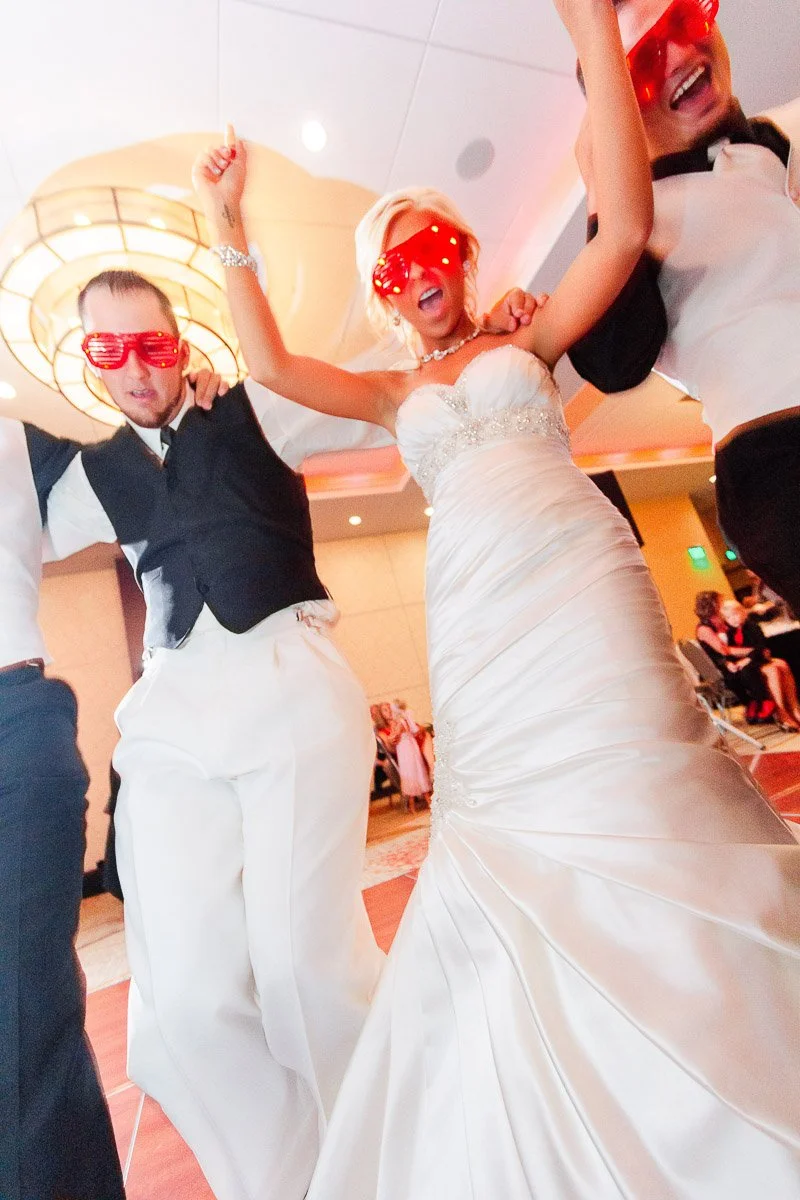 A bride and two men, all wearing red heart-shaped glasses, joyfully dance at a wedding reception. The atmosphere is lively and festive.