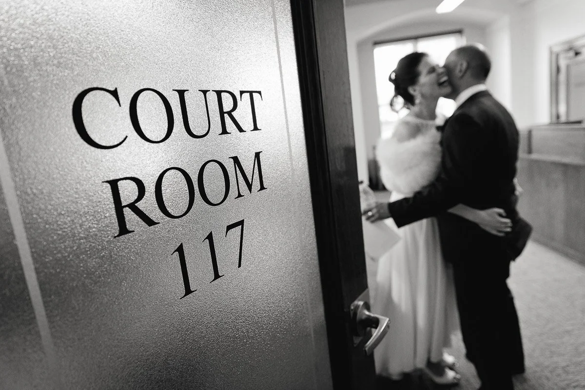 A couple embraces joyfully behind a frosted glass door marked "Court Room 117," conveying a sense of love and celebration within a formal setting.