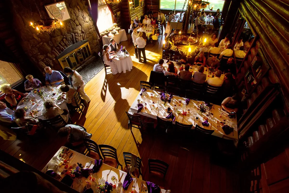 A cozy dining room with wooden floors is filled with round and rectangular tables set for a wedding. Warm lighting and stone walls create an inviting atmosphere.