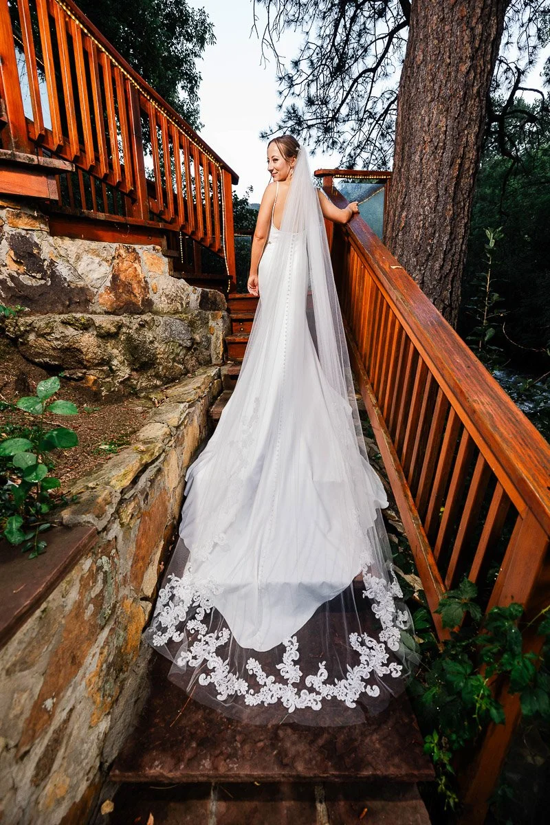 Bride in a long white gown and veil with lace detail ascends stone steps lined with wooden railings, surrounded by lush greenery and trees.
