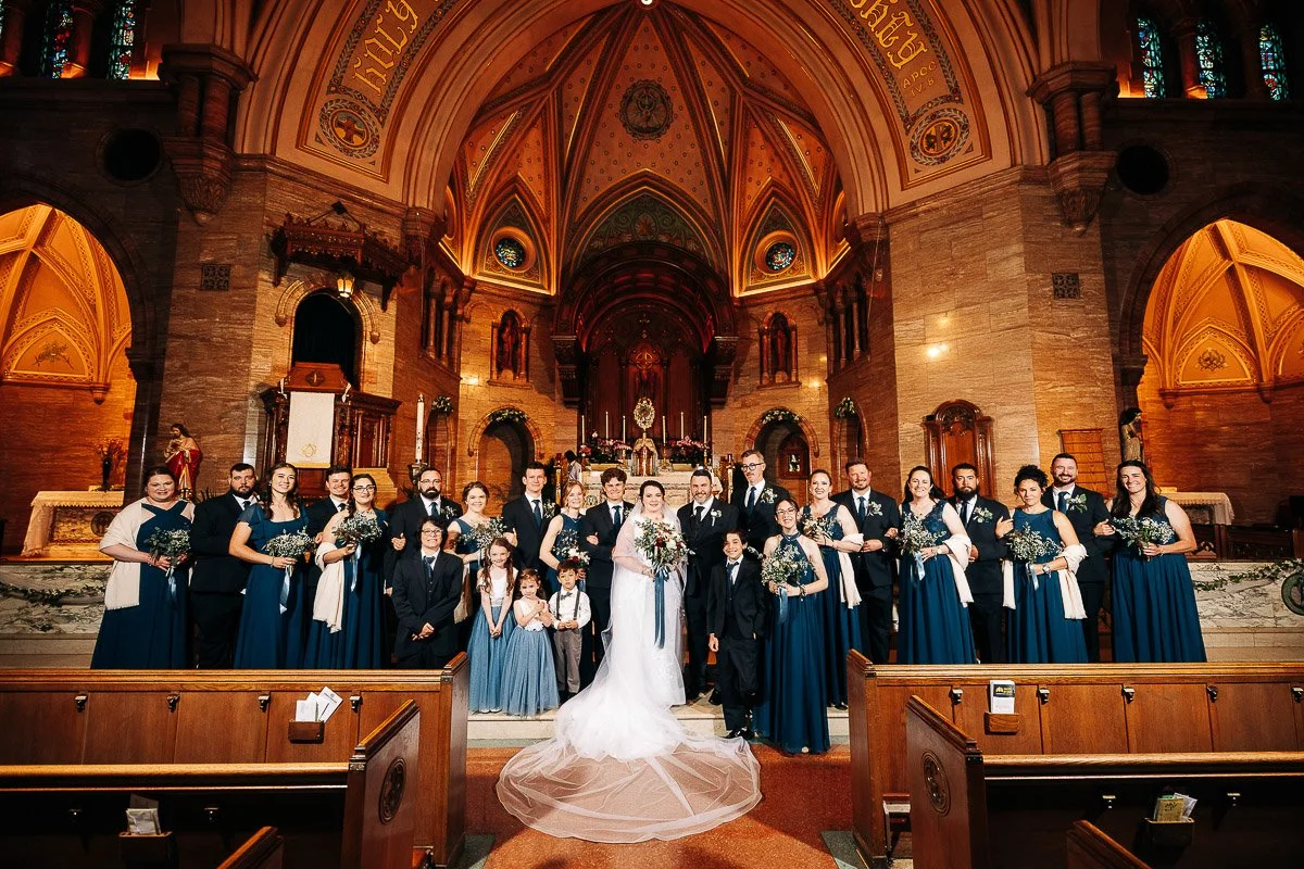 A wedding party poses inside a grand church with ornate arches and warm lighting. The bride and groom stand at the center, surrounded by joyful guests.