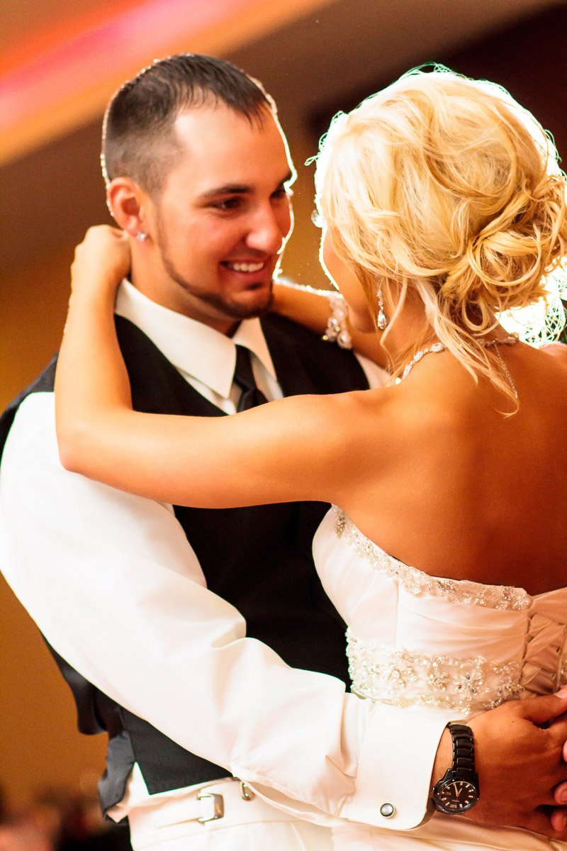 A joyful couple dances closely at a wedding, with the groom in a black vest and tie, and the bride in a white gown, both smiling warmly.