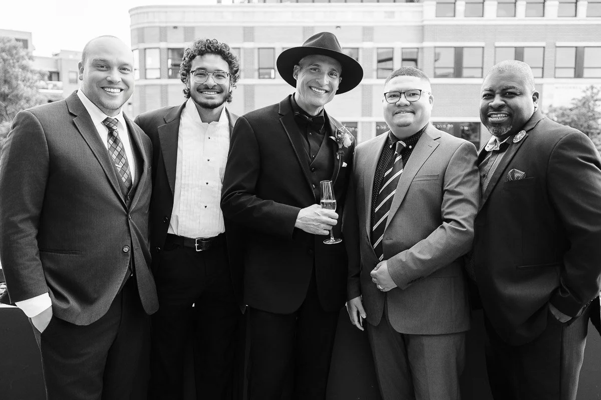 A group of five men in formal wear stand closely together on a balcony, smiling at the camera. The mood is joyful and celebratory, in a city setting.
