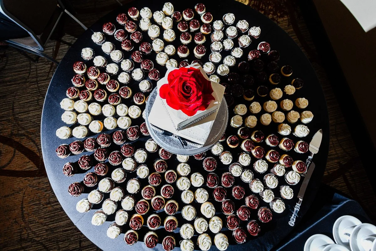 A round table with a central two-tier cake topped by a red rose, surrounded by concentric circles of white and dark chocolate cupcakes, creating a festive display.