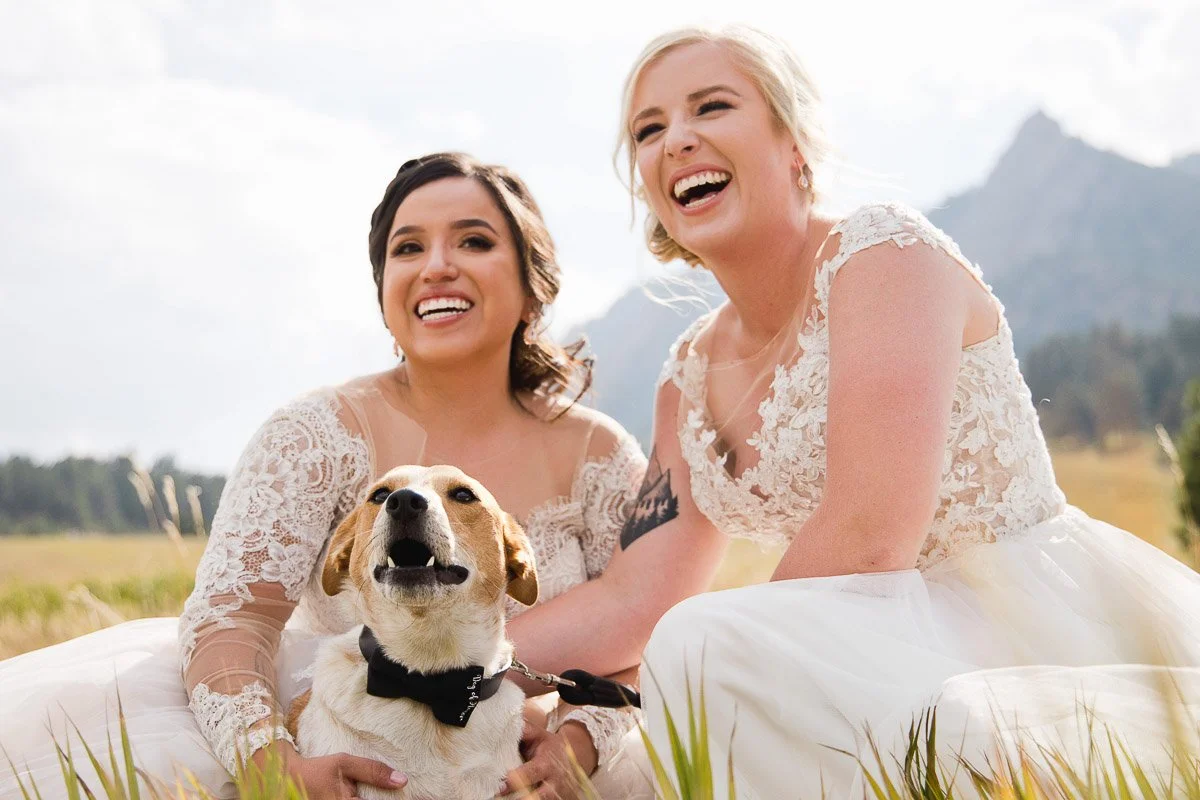 Two smiling women in lace wedding dresses sit on grass in Chautauqua Park with a happy dog captured by Boulder Wedding Photographer tomKphoto