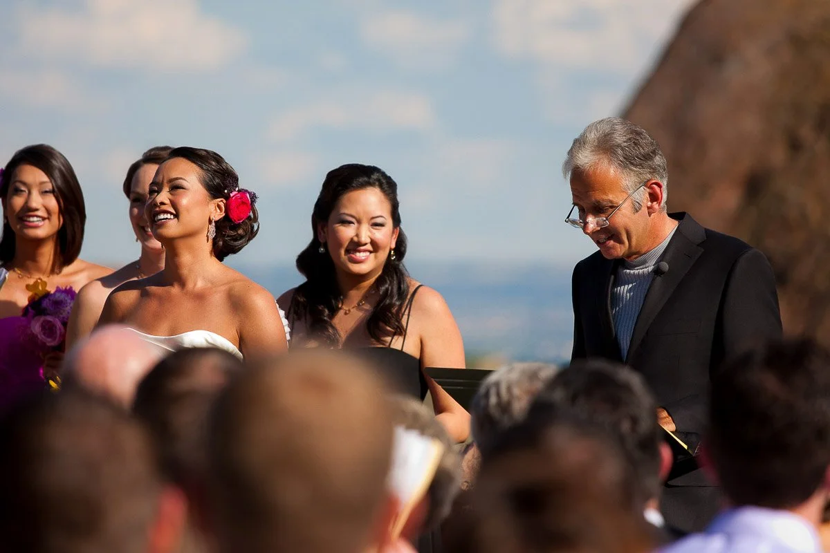 Bride laughing joyfully with bridesmaids and an officiant during an outdoor wedding ceremony. Sunny day with a scenic background, conveying happiness.