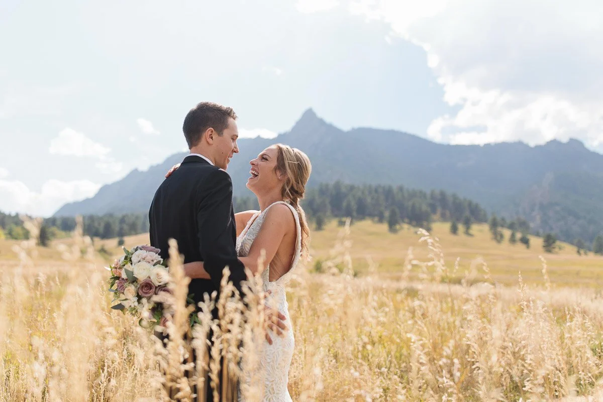 A bride and groom share a joyful moment in a sunlit field with mountains in the background. She holds a bouquet, and both are dressed elegantly.