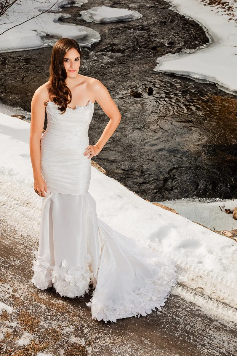 A woman in a strapless white wedding gown stands confidently by a snowy riverside. Her long hair is styled neatly, and the scene conveys elegance.