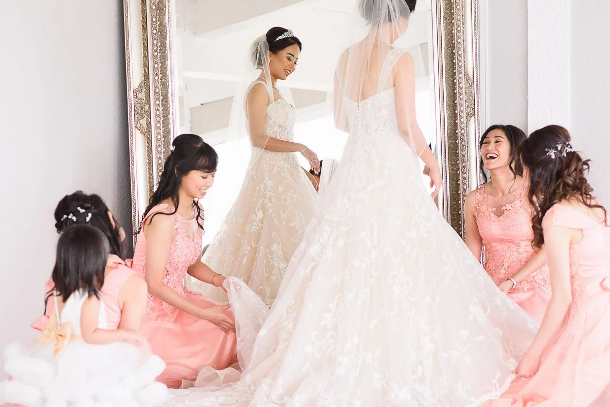 Bride in an elegant lace gown stands before a mirror, smiling, surrounded by joyful bridesmaids in pink dresses, adjusting her train.