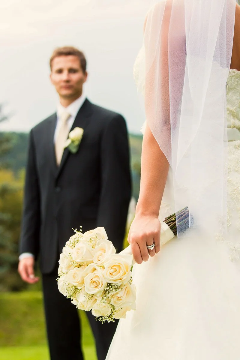 Bride holding white roses, focuses on bouquet and wedding dress detail. Groom in suit, blurred background. Outdoor setting, serene mood.