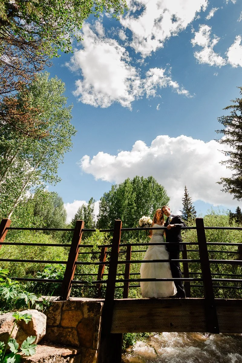 A bride and groom embrace on a wooden bridge amidst lush greenery under a bright sky with fluffy clouds, conveying joy and romance.
