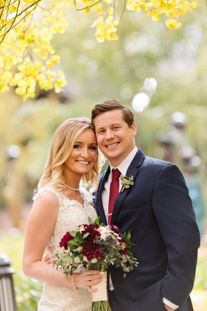 A couple smiling warmly on their wedding day. The bride holds a bouquet of burgundy and white flowers, while yellow leaves frame the joyful scene.