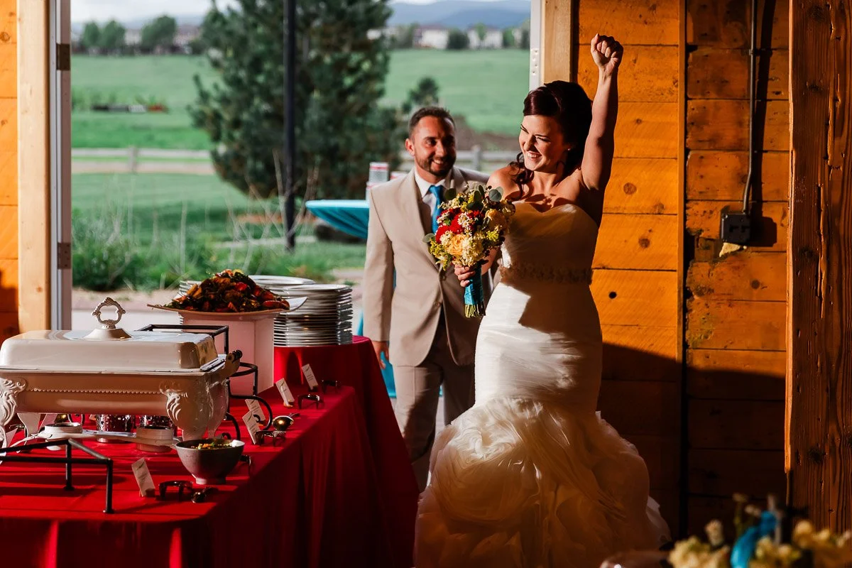 Bride and groom enter a rustic venue, the bride raises her fist joyfully holding a colorful bouquet. A red-clothed buffet table is to the side.