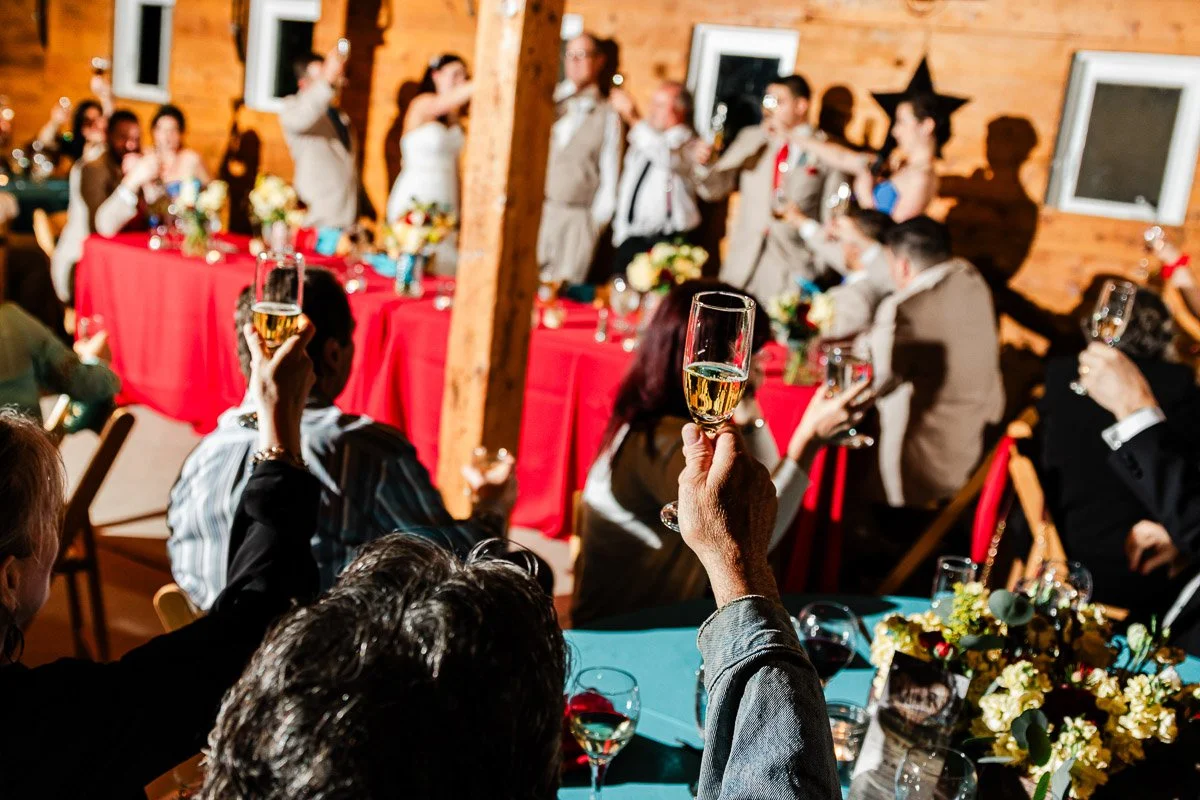 Guests at a festive event raise glasses in a toast. People are seated at tables with vibrant red tablecloths and floral centerpieces, creating a joyful atmosphere.