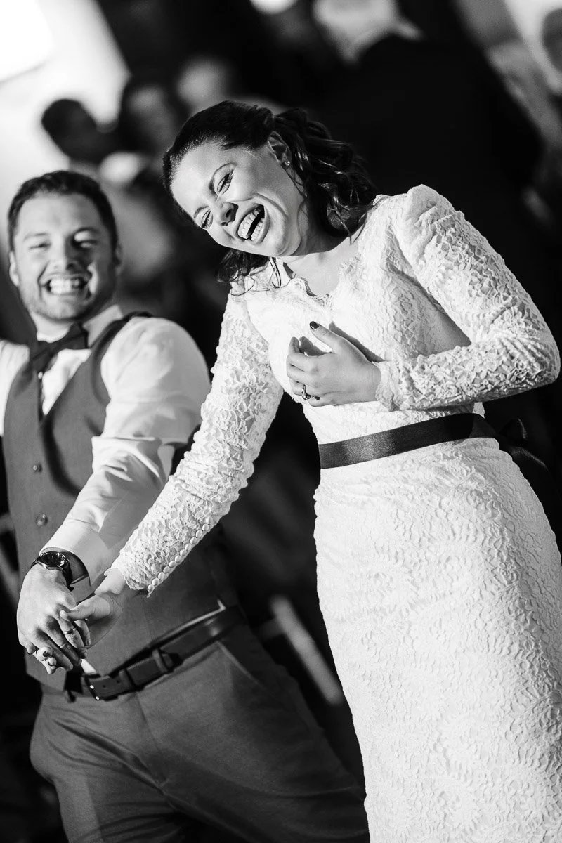 A joyful couple, in formal attire, dances and laughs together. The woman wears a lace dress, and the man is in a vest. The mood is celebratory and lively.