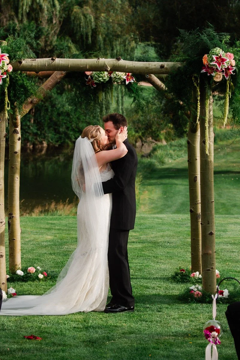 A newlywed couple shares a kiss under a floral-decorated wooden arch in a garden setting, exuding romance and joy with vibrant greenery around.