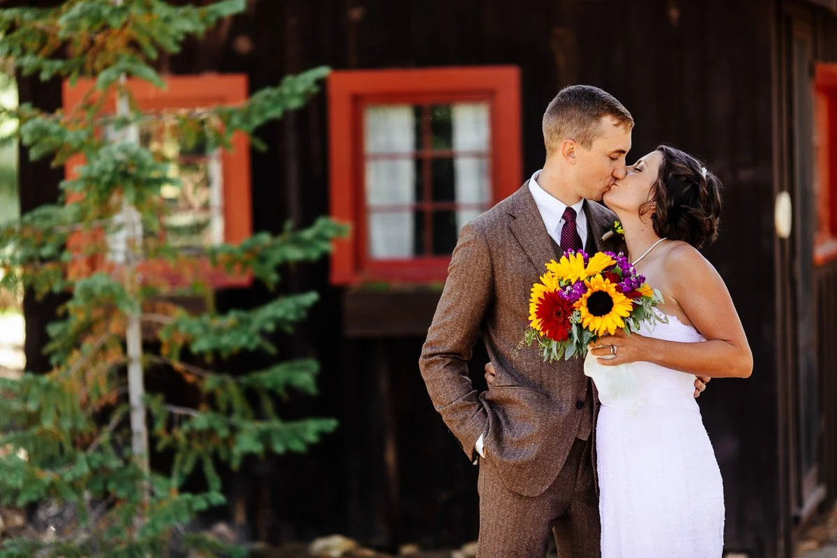Couple kissing at a wedding, with the groom in a brown suit and bride in a white dress holding a bouquet of vibrant sunflowers. Rustic cabin backdrop.