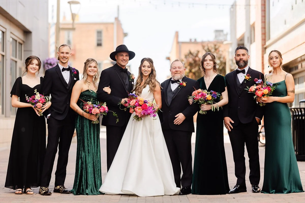 A wedding party poses outdoors on a city street. The bride in a white gown is surrounded by bridesmaids in dark dresses and groomsmen in suits. Everyone holds vibrant bouquets, conveying joy and celebration.
