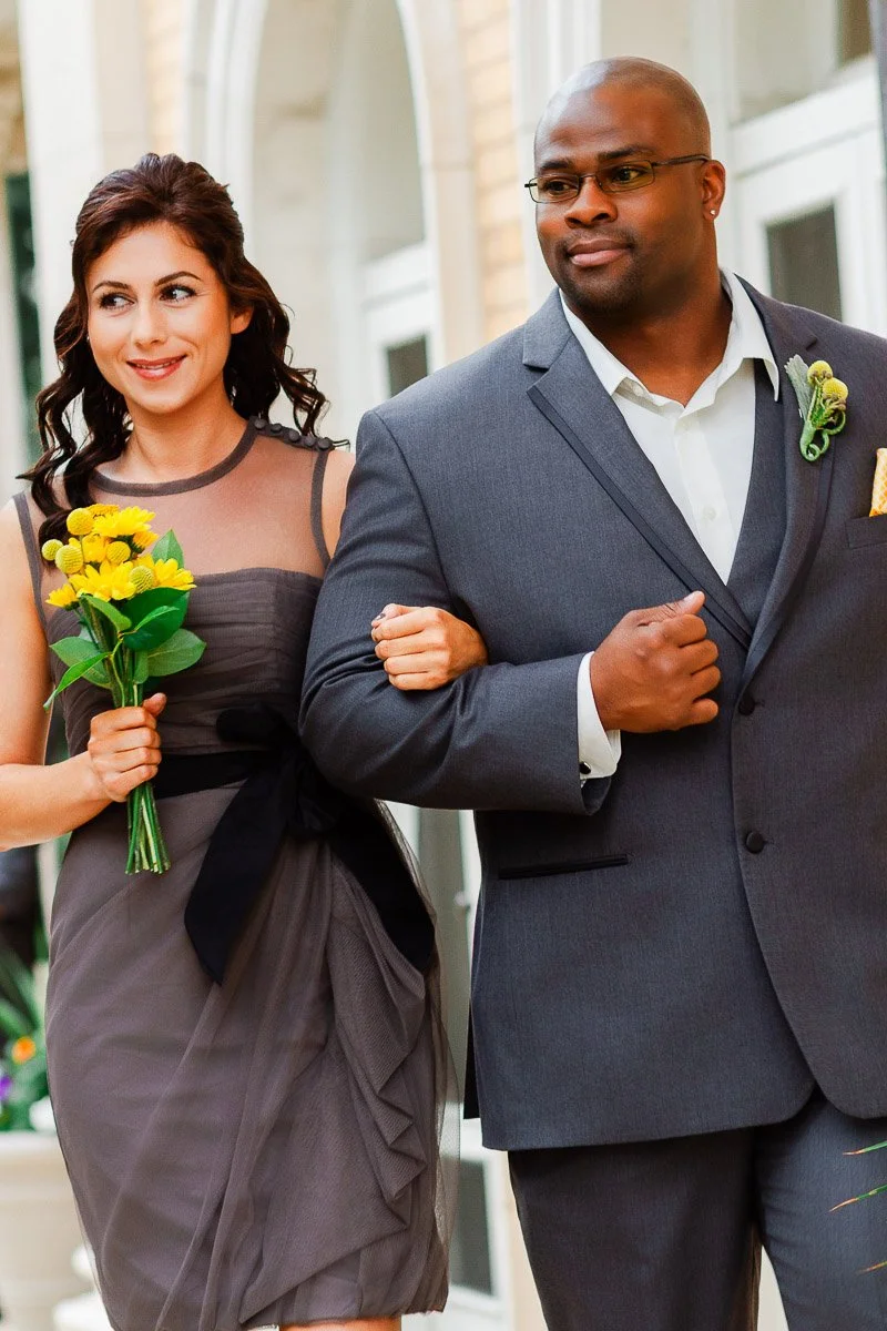 A woman in a gray dress holding yellow flowers walks arm-in-arm with a man in a gray suit. They appear happy, with a wedding ambiance.